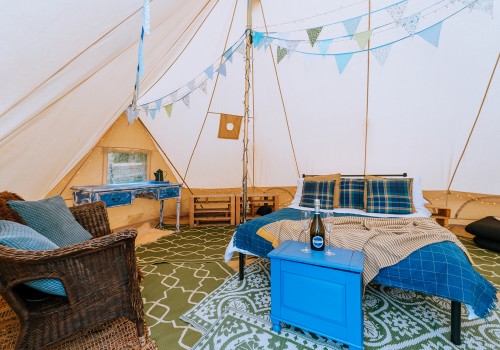 The inside of a cotton canvas bell tent, a double bed is in the centre of the tent dressed with bed linen, up-cycled furniture is on display and woven accent chair in the corner. Green pattern rugs are used as the flooring base inside the tent,
Layers of pale blue and light green bunting hang between tent poles.