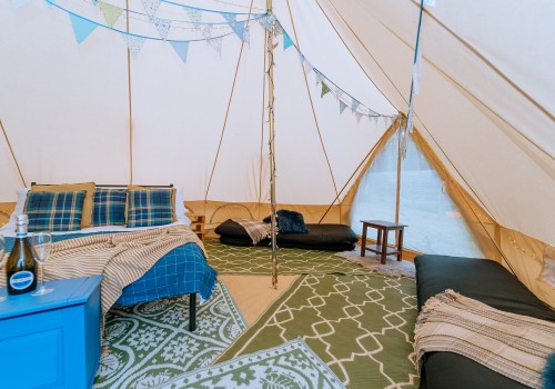 A colourful interior of the bell tent with 4 patterned rugs underfoot. Cotton Canvas tent sides meet the high emperor ceiling. Pale blue bunting and striped blankets placed inside the tent. Four floor mattresses, two are stacked together on each side of the tent. 
A dressed double bed with additional blankets and pillows.